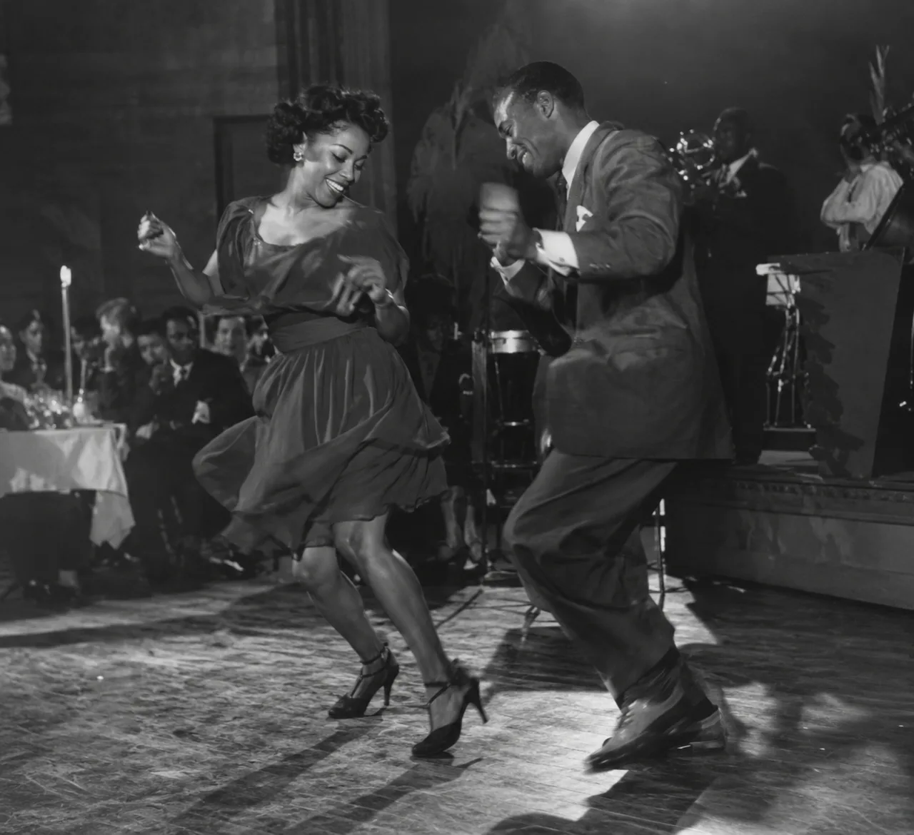 Dos bailarines improvisan pasos de jazz uno junto al otro frente a una banda